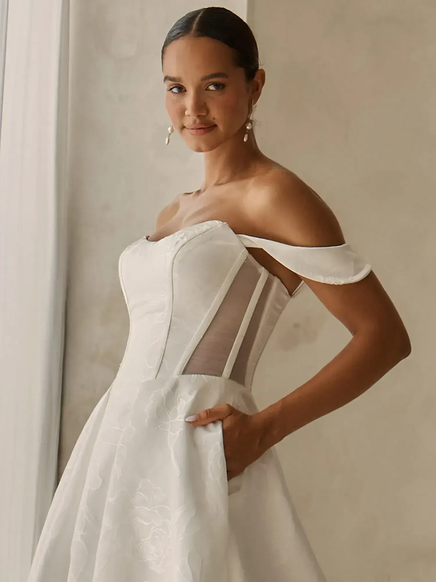 A woman wearing an elegant off-the-shoulder white dress with a fitted bodice and a full skirt, posing confidently.