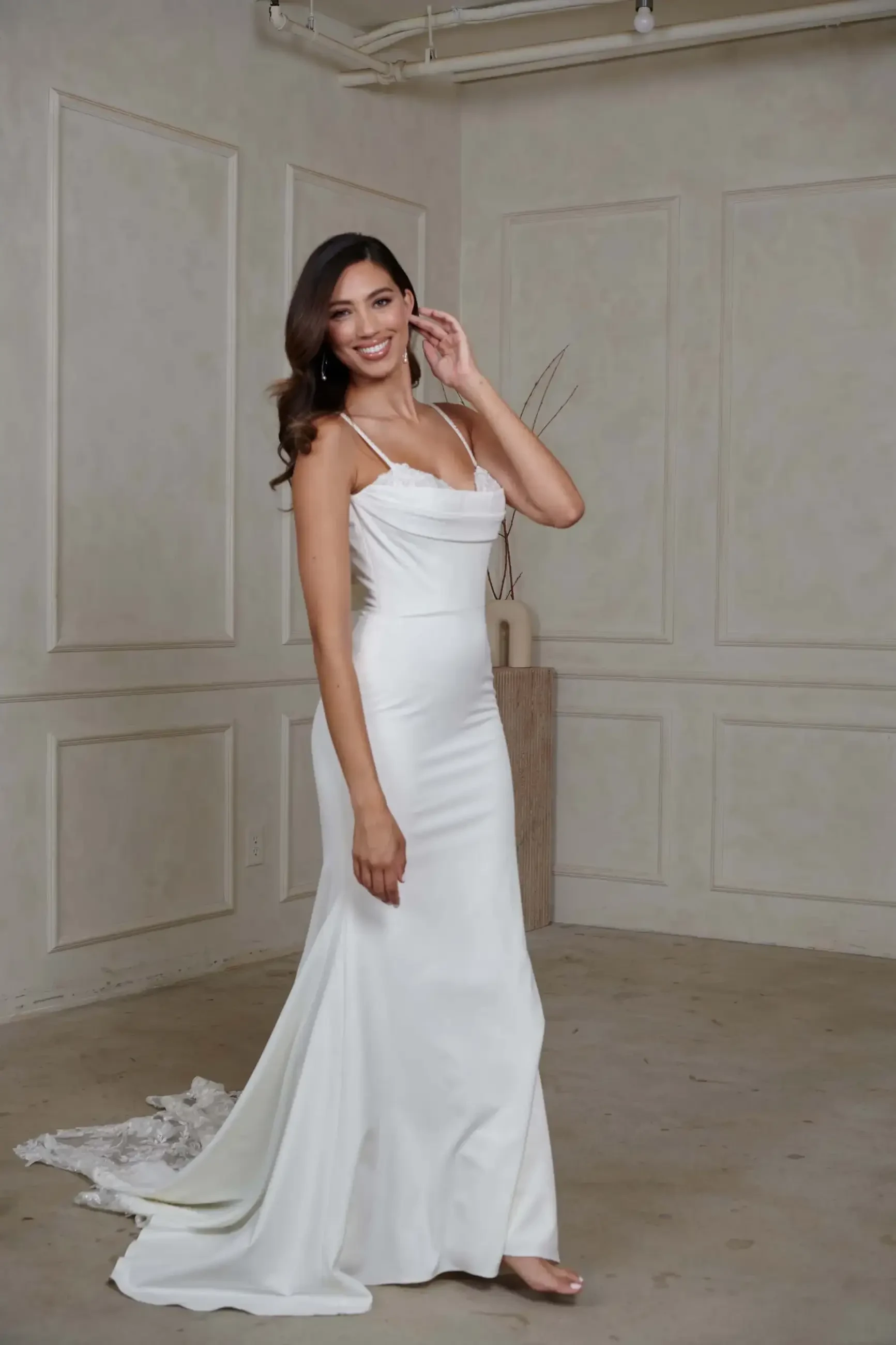 A woman wearing a sleek white wedding gown with straps stands in a minimalist room, smiling warmly. The elegant dress has a small train.