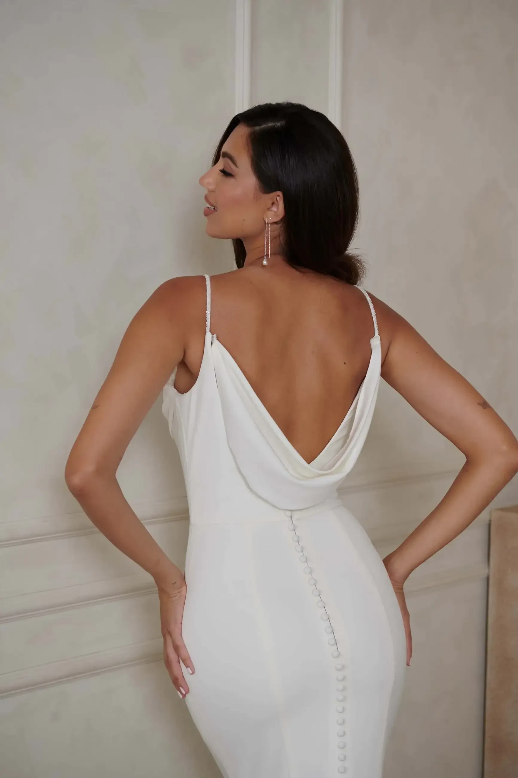 Woman in an elegant, backless white gown with button details, standing against a light-colored wall. She exudes a confident, serene demeanor.