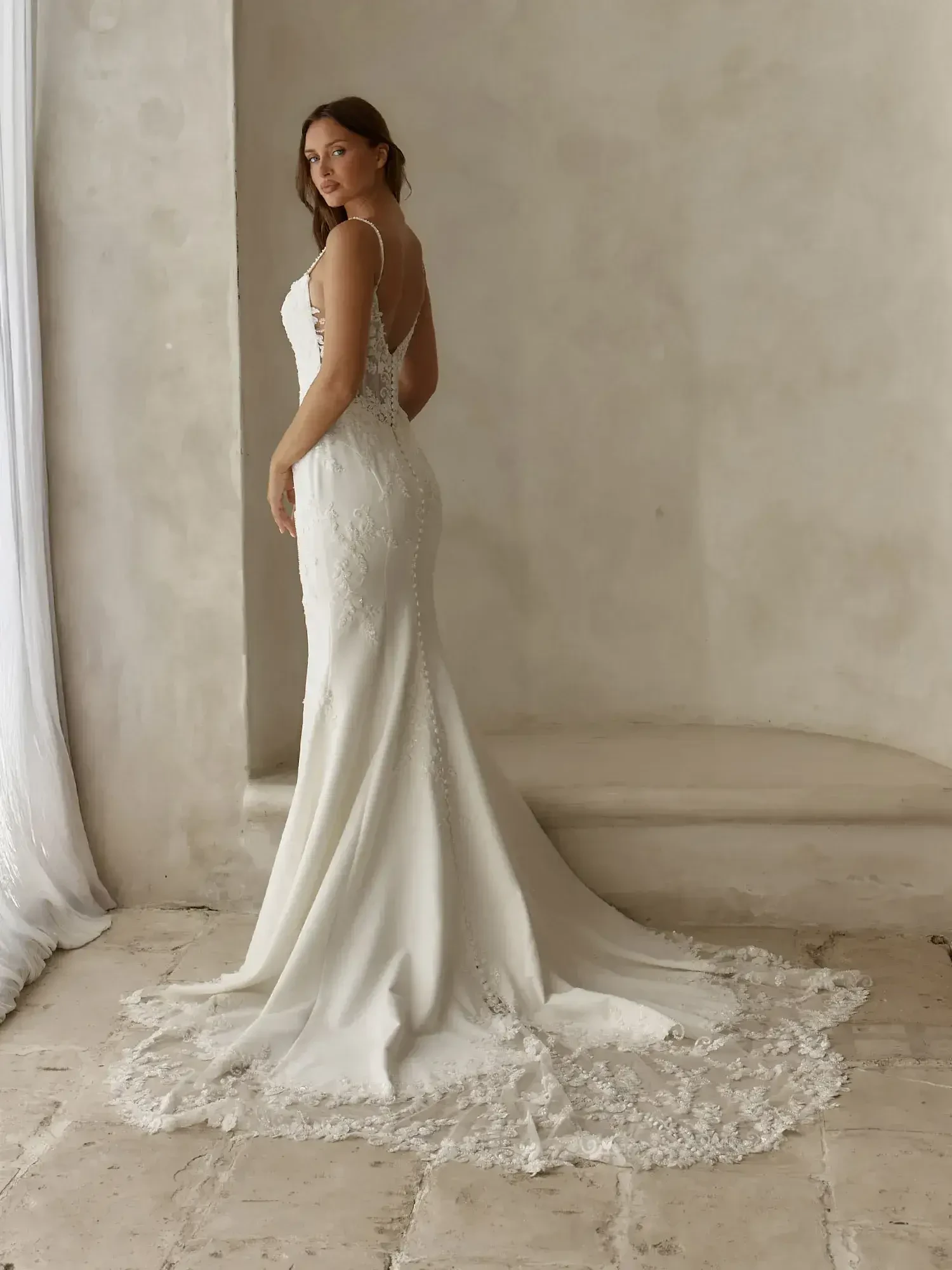 A woman in a fitted white gown with lace details, standing sideways and looking back over her shoulder. The gown has spaghetti straps and a long train.