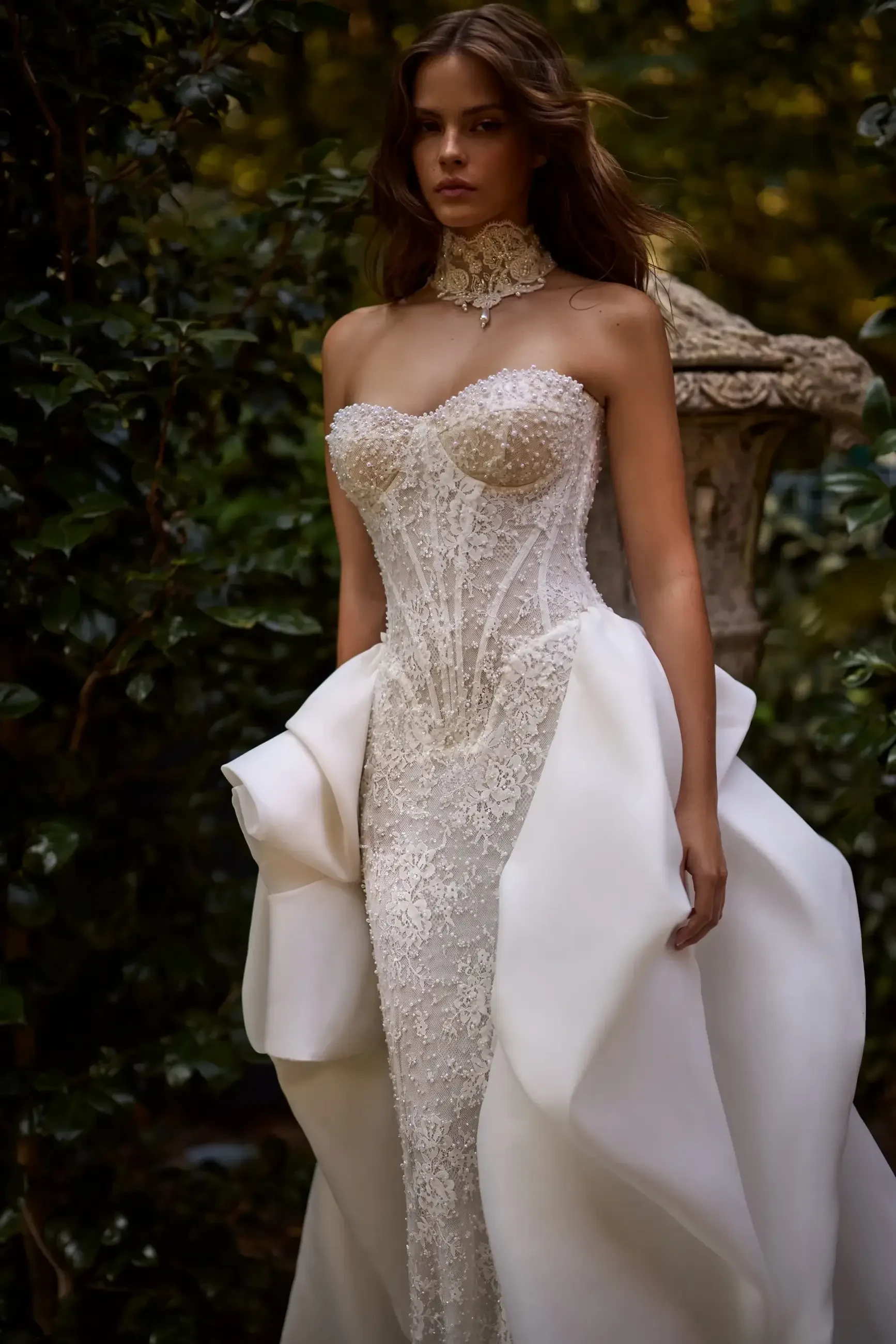 A model wearing an elegant, fitted wedding dress with intricate lace detailing and a flowing white train, walking outdoors among greenery.