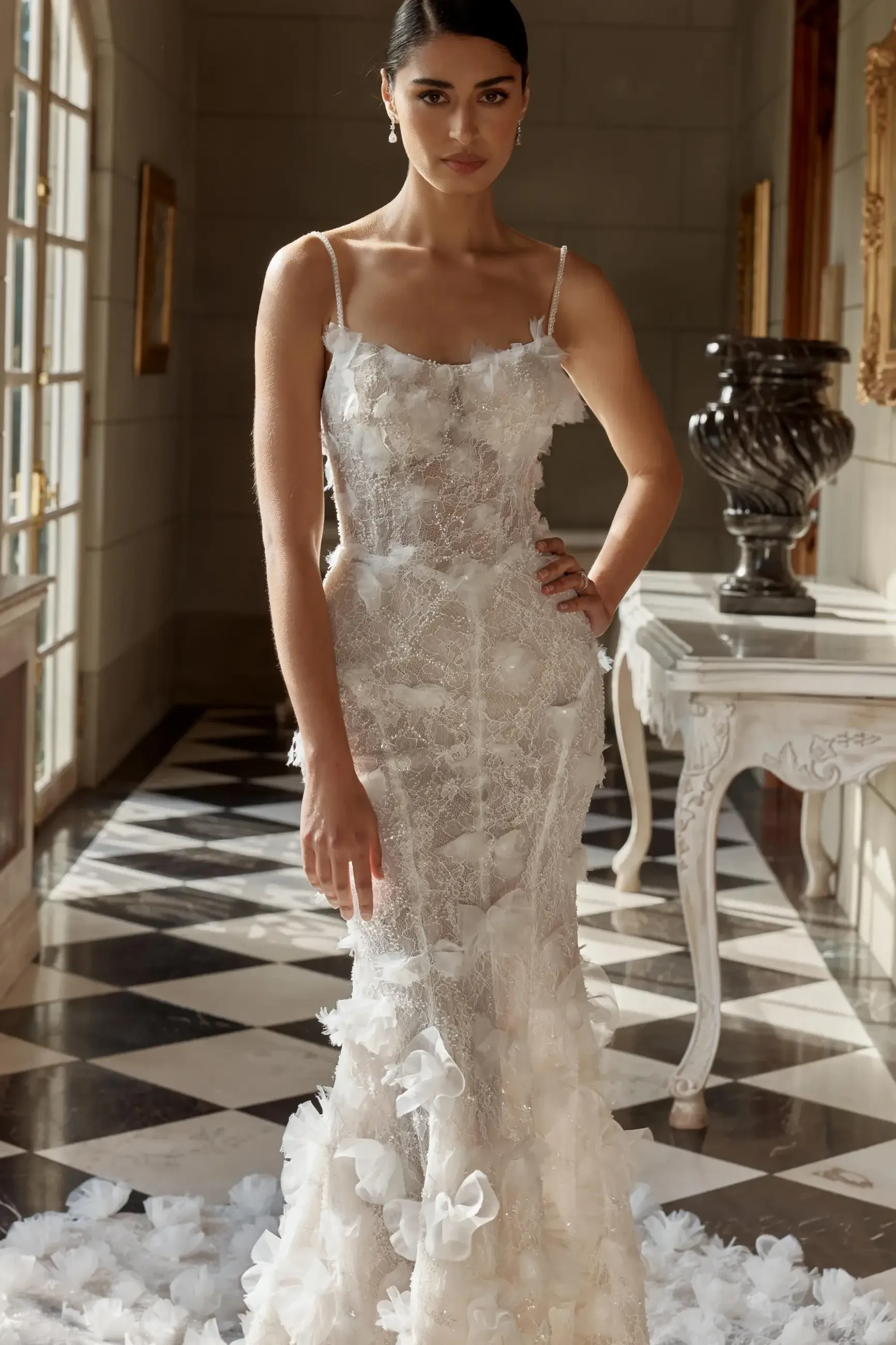 A woman wearing a delicate, fitted white gown adorned with floral details, standing elegantly in a well-lit room with checkered flooring.