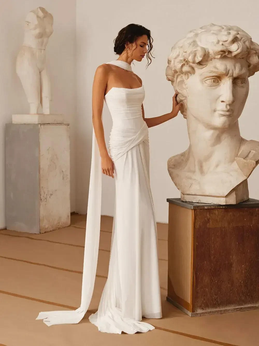 A woman in a white strapless gown stands beside a marble statue of a classical figure.