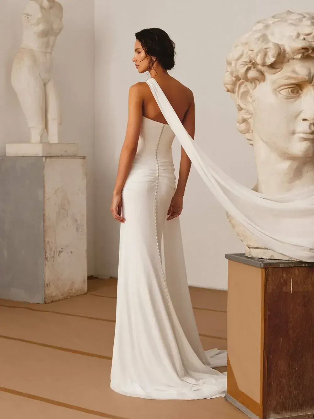 A woman in a sleek white gown stands with her back to the camera, draped fabric flowing behind her, in a minimalist setting with sculptures in the background.