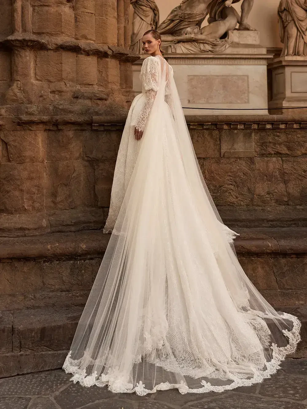 A model in a flowing white wedding dress with a long train, posed against a grand stone backdrop.