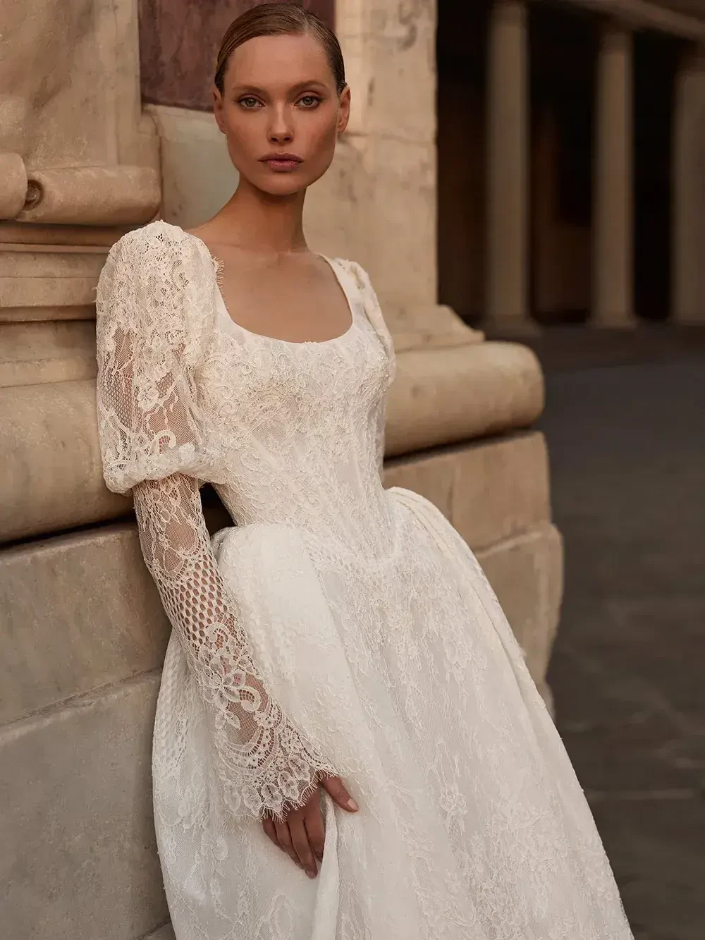 A model in a white dress with lace sleeves stands against a stone wall.