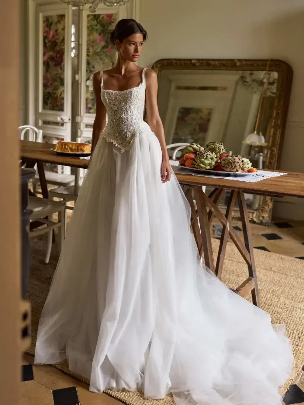 A woman in an elegant white wedding dress with a lace bodice and flowing skirt stands by a rustic wooden table adorned with fruits and bread, in a vintage room.