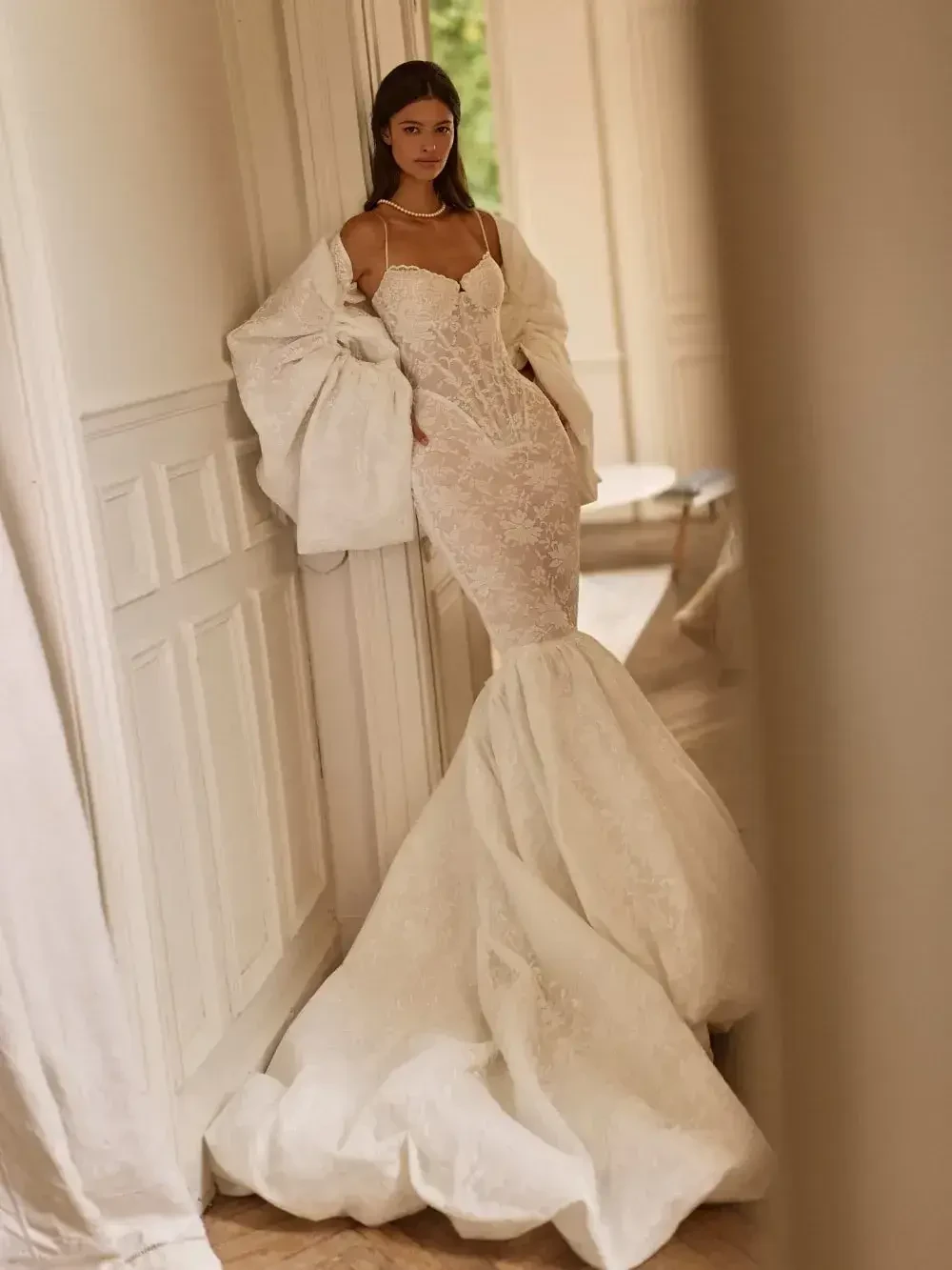 A woman in a fitted white wedding dress with a mermaid silhouette and a dramatic train, wearing a light shawl, standing elegantly in an indoor setting.