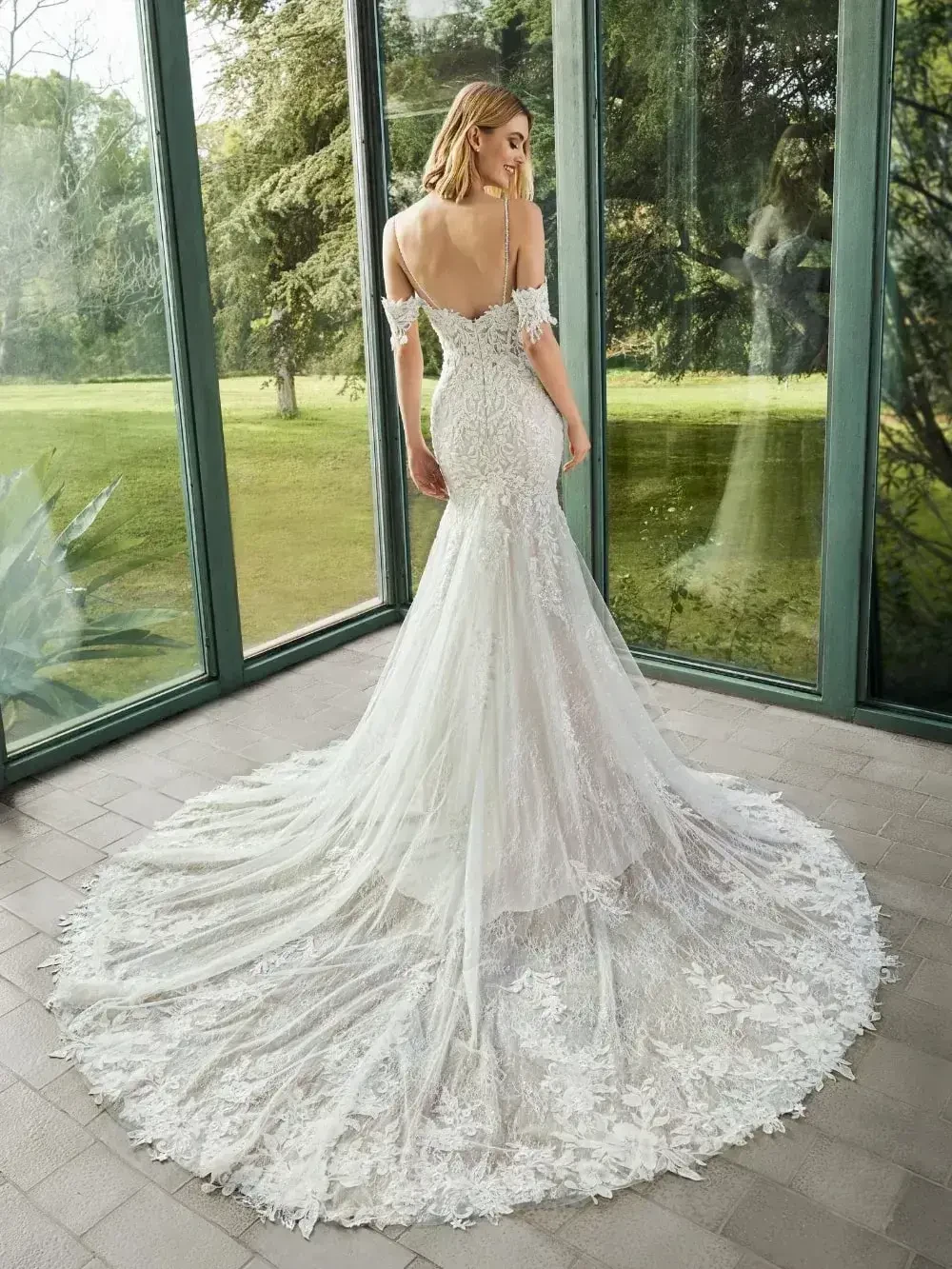 A bride wearing an elegant wedding dress with intricate lace details, standing with her back to the viewer. The dress has a long train and off-the-shoulder sleeves. She is in a sunlit room surrounded by large windows, with a view of greenery outside.