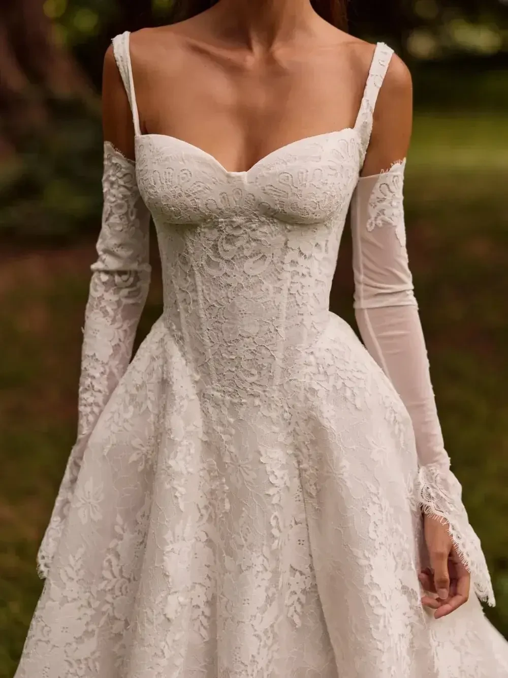 A close-up of a woman in an elegant white lace wedding dress with off-the-shoulder sleeves. The dress features intricate floral patterns and exudes a romantic, timeless feel. The background is a blurred, outdoor setting.