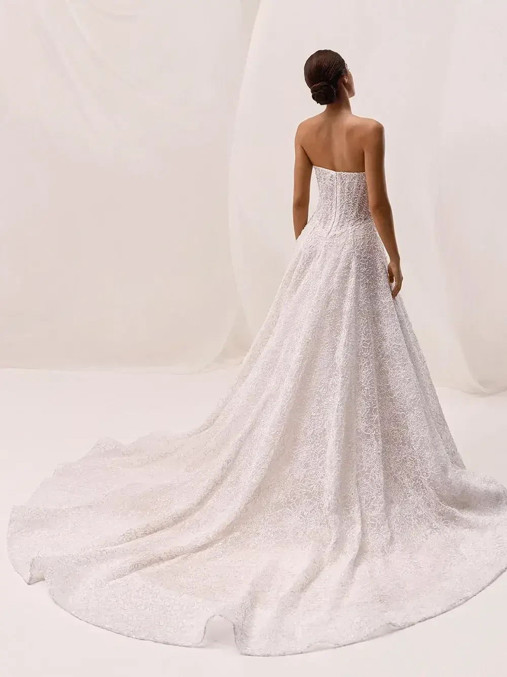 A woman wearing a strapless white gown, viewed from the back, with a long train extending behind her against a pale background.