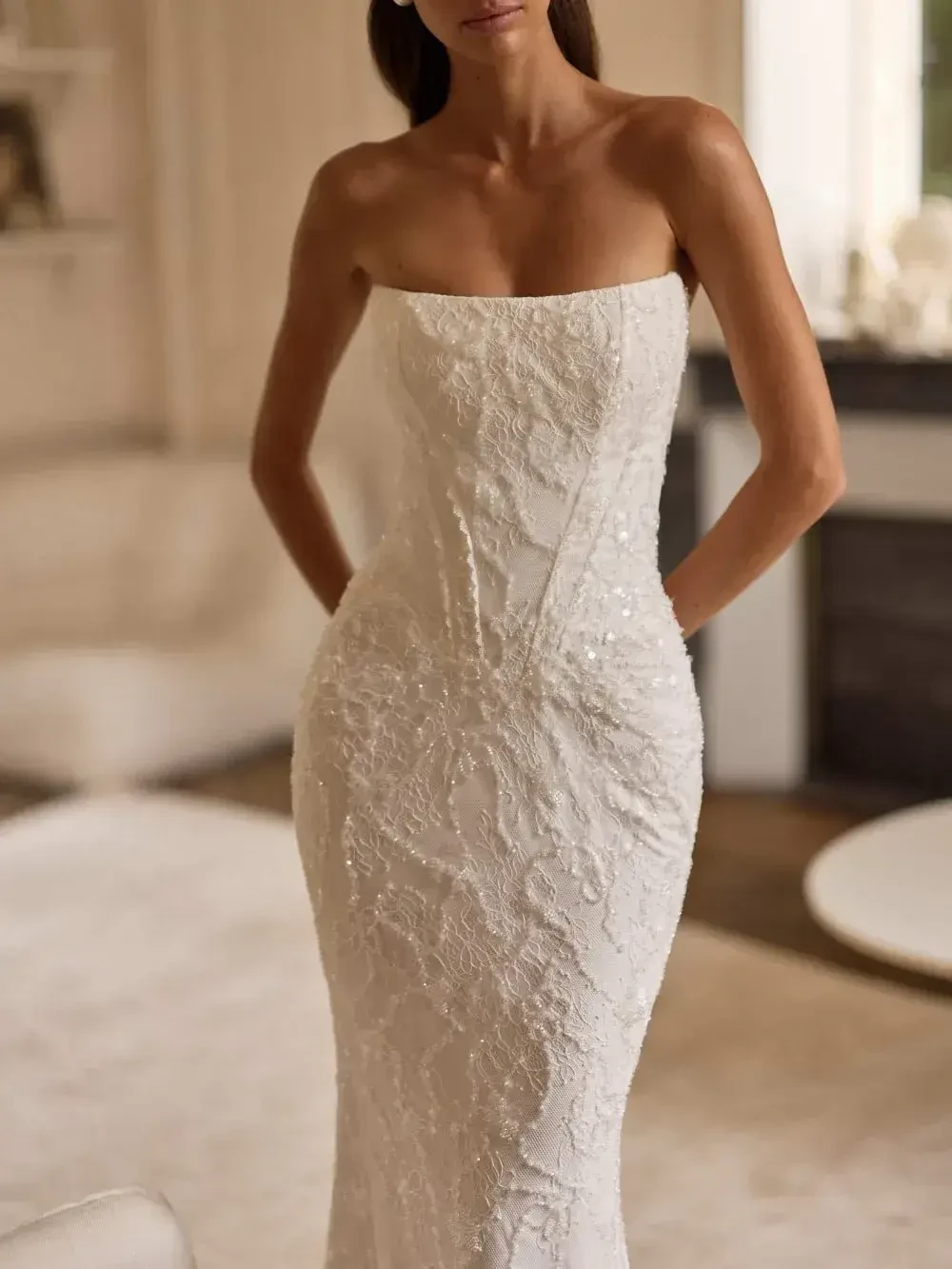 A woman wearing a strapless white dress with intricate detailing, standing in a well-lit room with light-colored decor.