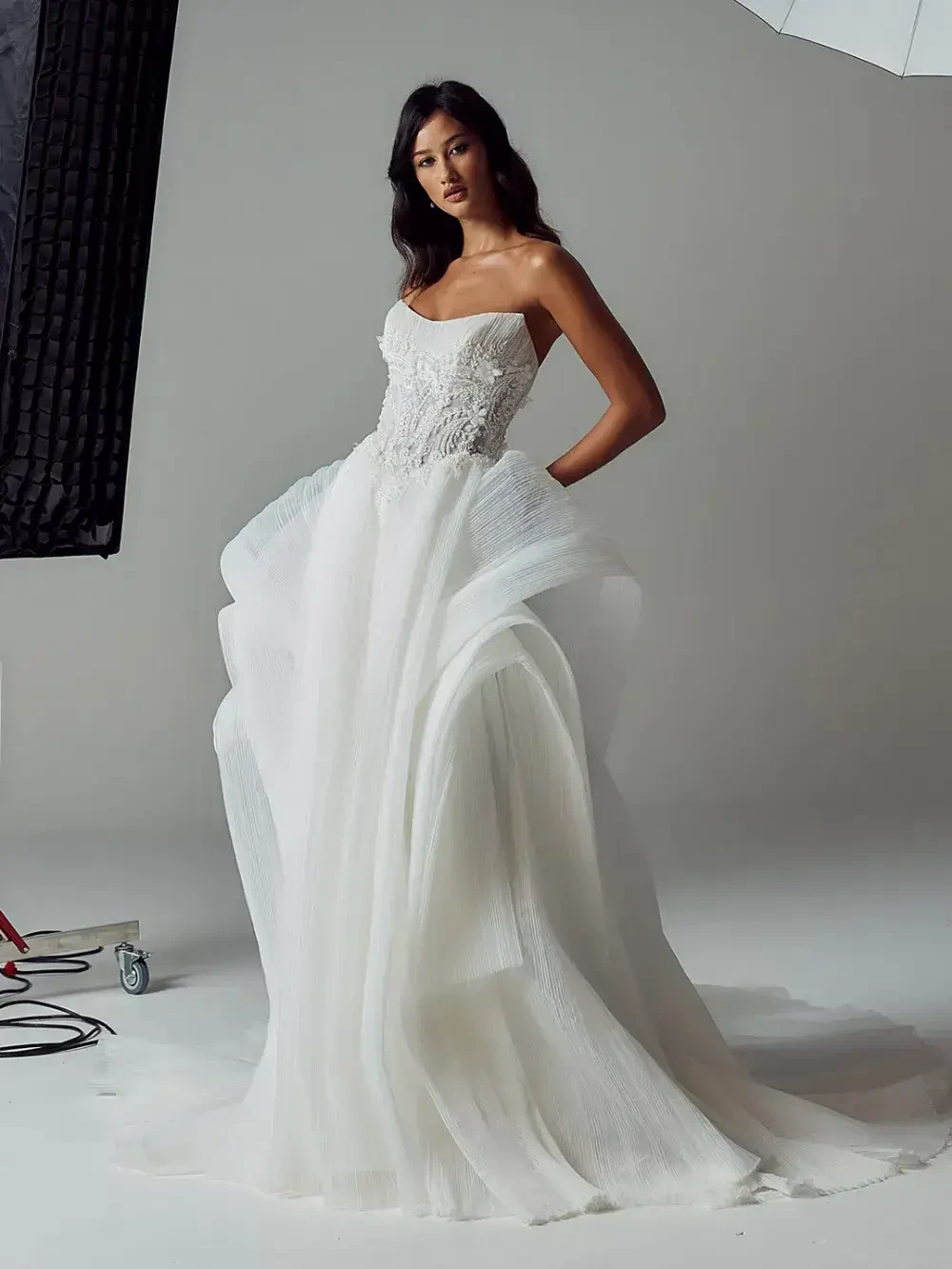A model wearing a strapless, white wedding dress with a detailed bodice and flowing skirt, posing in a studio setting.