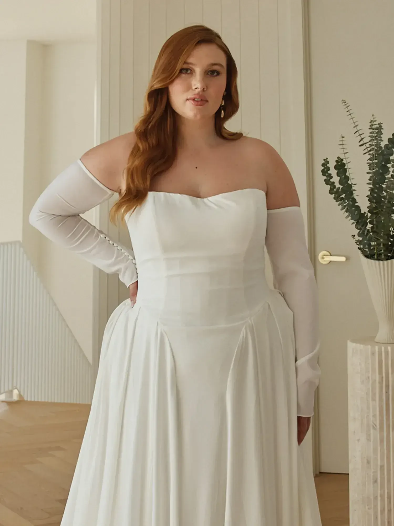 A woman wearing a white off-the-shoulder dress stands in a room with light wood flooring and neutral-toned walls, striking a pose with one hand on her hip.