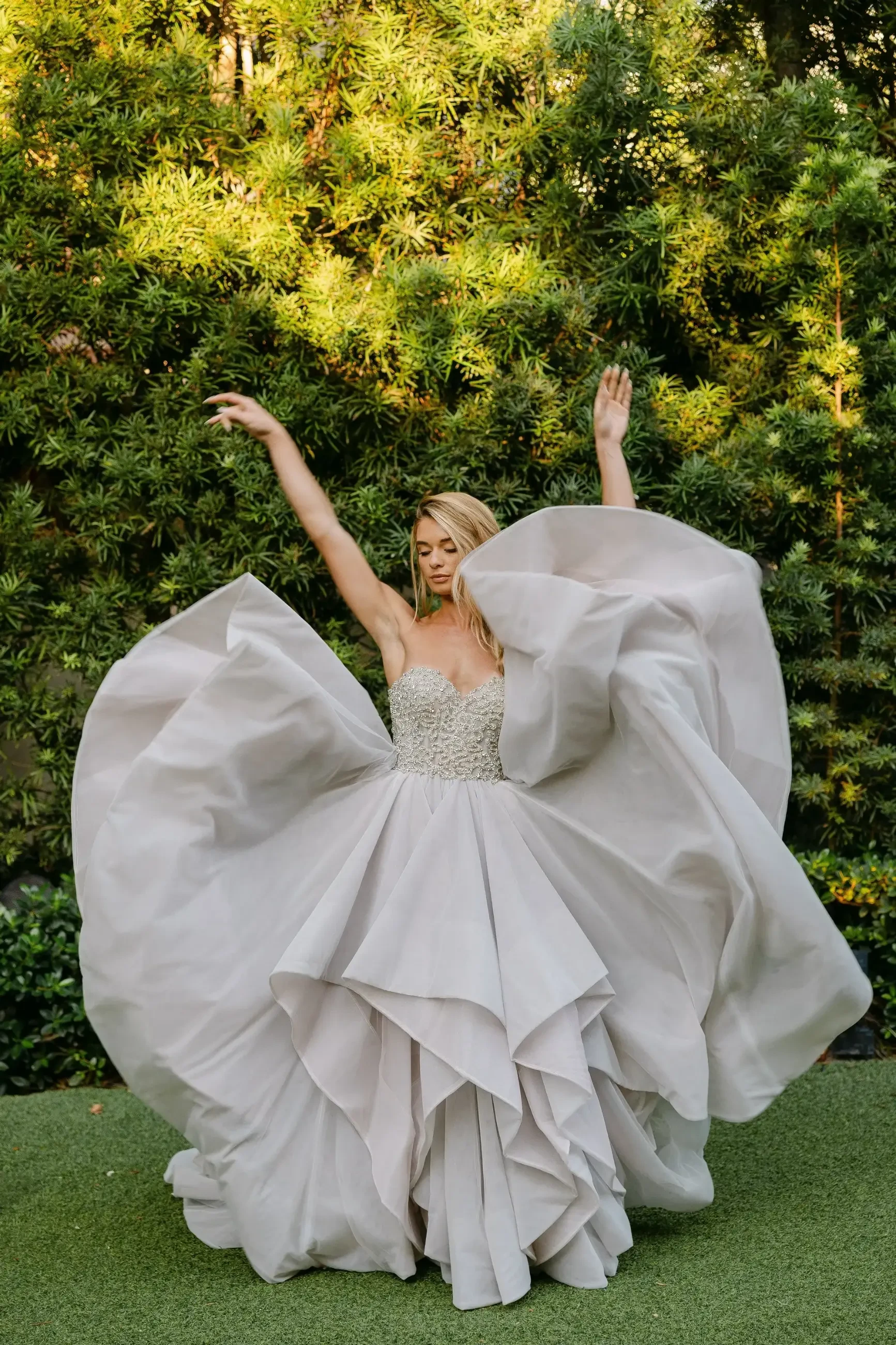A woman in a flowing, elegant gray gown is dancing against a backdrop of lush greenery. Her arms are raised gracefully, and the dress has large layers that create a dramatic effect.