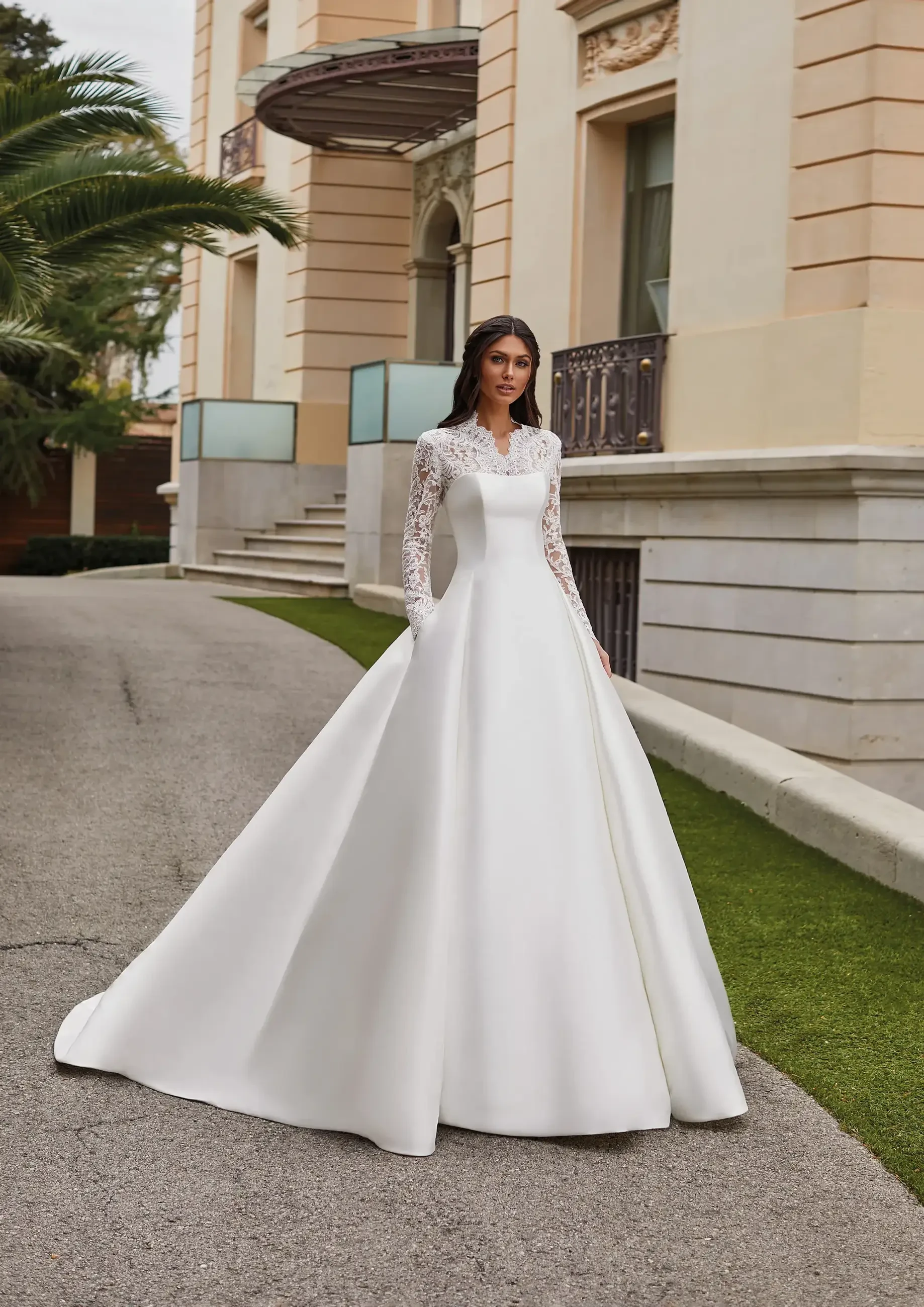 A woman in a long, white wedding dress with lace sleeves standing on a pathway beside a building.