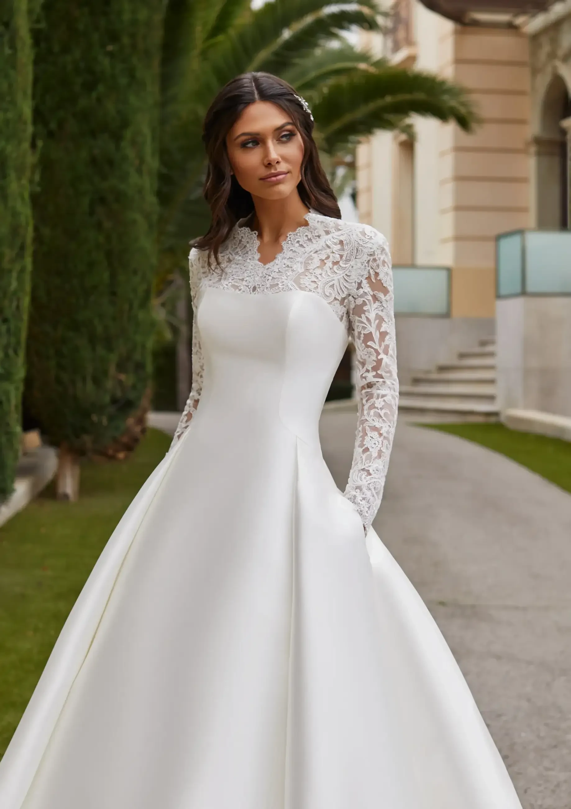 Bride in elegant white gown with lace sleeves and pockets, stands on a garden path. Background features lush greenery and a grand building.