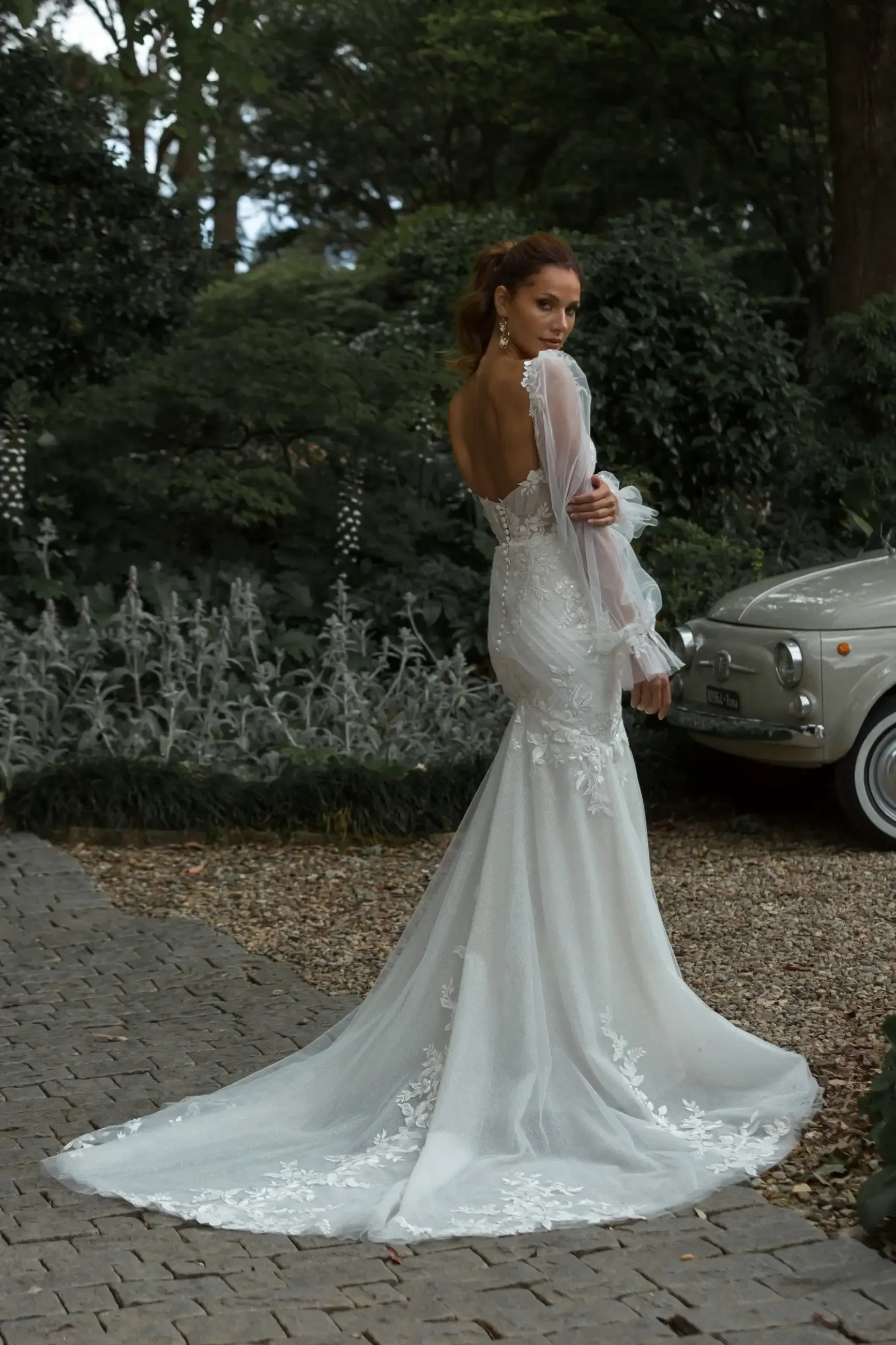 A woman in a bridal gown stands outdoors with her back to the camera, showcasing intricate lace details and a flowing train. She is surrounded by greenery and a vintage car is parked nearby.