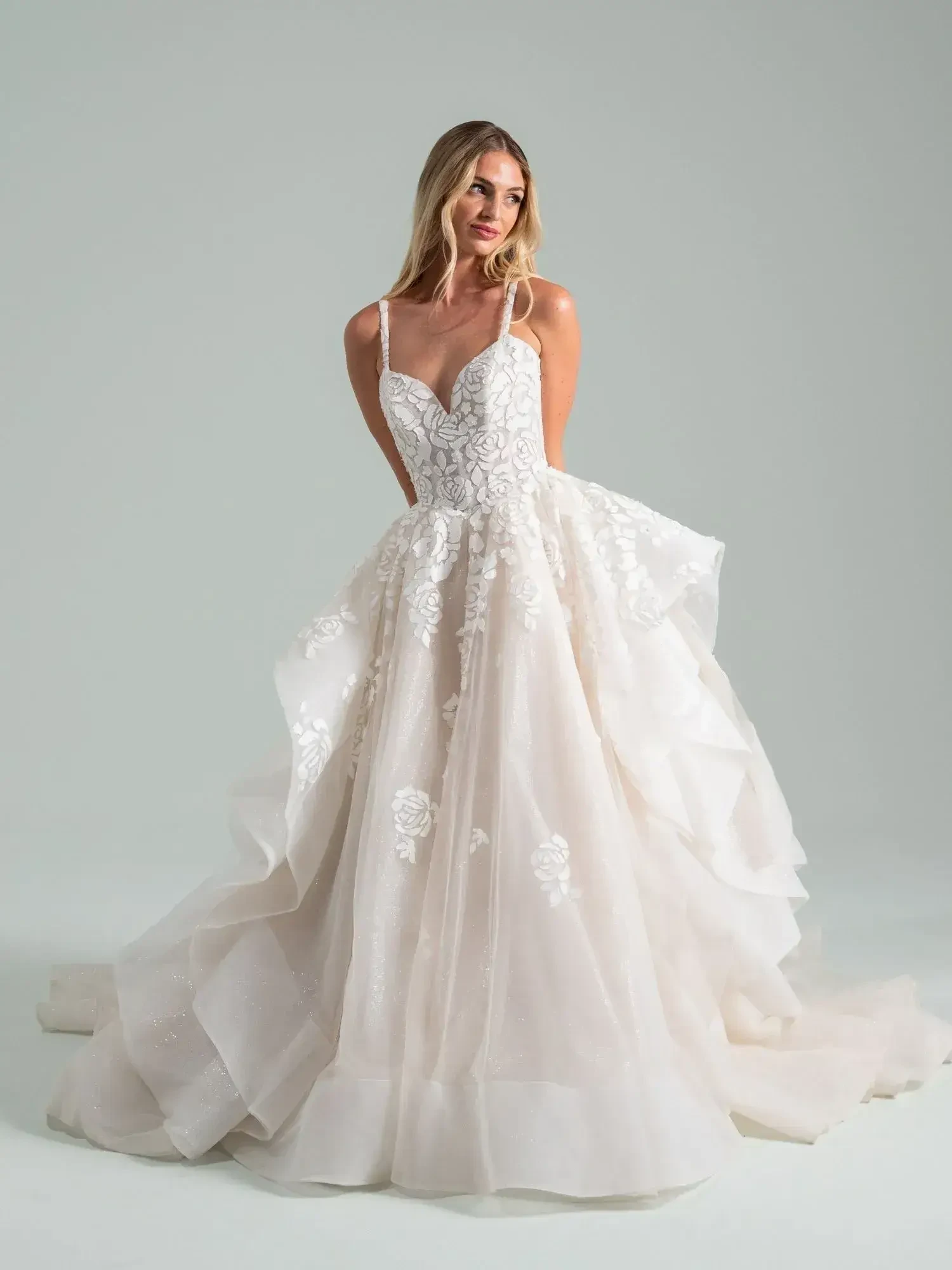 A woman in an elegant ivory gown poses gracefully. The dress features floral lace details, a fitted bodice, and a voluminous skirt, conveying a romantic tone.