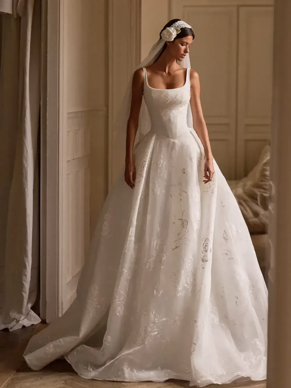A woman wearing a wedding dress with a fitted bodice and a flowing skirt, standing in an elegant indoor setting. She has a veil and floral hair accessory.