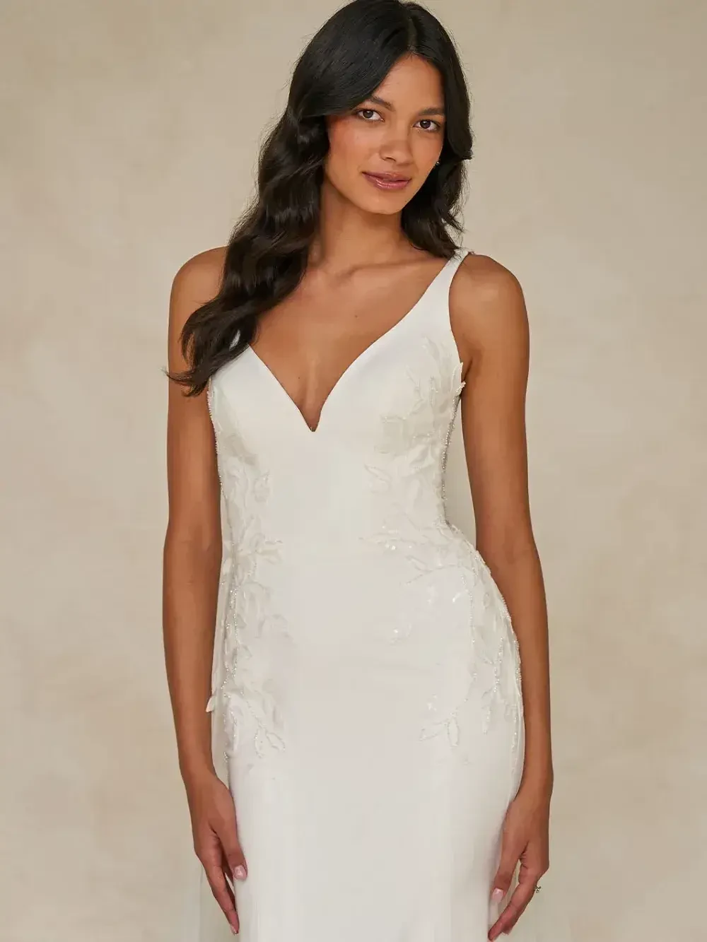 A woman in a white wedding dress with a deep V-neckline stands against a beige background. Her long dark hair is styled in soft waves, conveying elegance.
