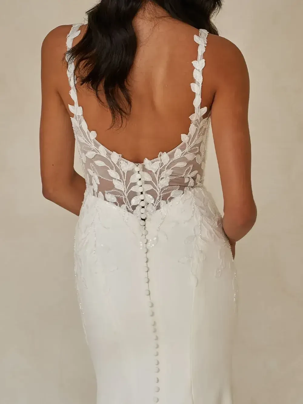 Bridal gown with a sheer lace back featuring leaf patterns and a row of buttons, worn by a woman with long dark hair cascading down her back.